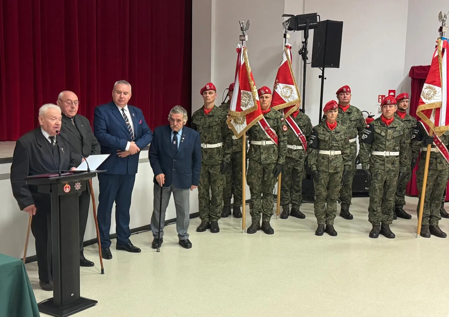 Grupa mężczyzn w garniturach i mundurach stojących w pomieszczeniu z jasną podłogą i bordową zasłoną w tle. Po lewej starszy mężczyzna przemawia przy czarnym mównicy, obok cztery osoby stojące z laskami. Po prawej osiem osób w mundurach wojskowych z czerwonymi beretami, trzy trzymają flagi.